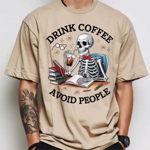 Puede incluir: Camiseta beige con un dise&ntilde;o de esqueleto con pajarita roja, bebiendo caf&eacute; y leyendo libros. El texto de la camiseta dice "DRINK COFFEE AVOID PEOPLE". El dise&ntilde;o incluye gafas, margaritas y un bol&iacute;grafo.