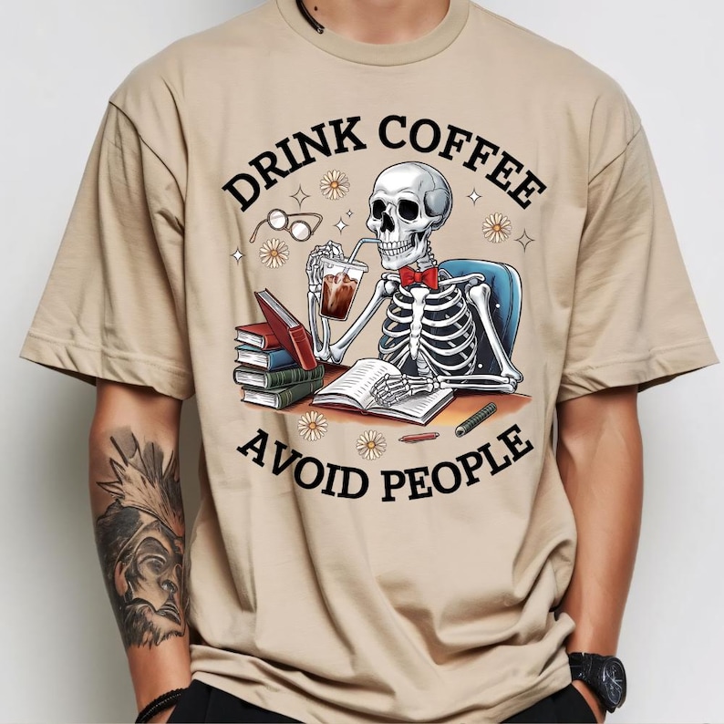 Puede incluir: Camiseta beige con un dise&ntilde;o de esqueleto con pajarita roja, bebiendo caf&eacute; y leyendo libros. El texto de la camiseta dice "DRINK COFFEE AVOID PEOPLE". El dise&ntilde;o incluye gafas, margaritas y un bol&iacute;grafo.