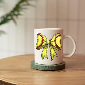 May include: White ceramic mug featuring a softball-themed bow design. The bow is yellow with red stitching, resembling a softball. The mug has a curved handle and sits on a green coaster. A decorative item for sports fans.