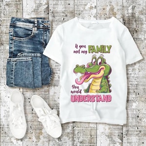 May include: White t-shirt with a cartoon alligator graphic and the text "If you met my FAMILY you would UNDERSTAND." The shirt is paired with blue jeans and white sneakers on a wooden surface.