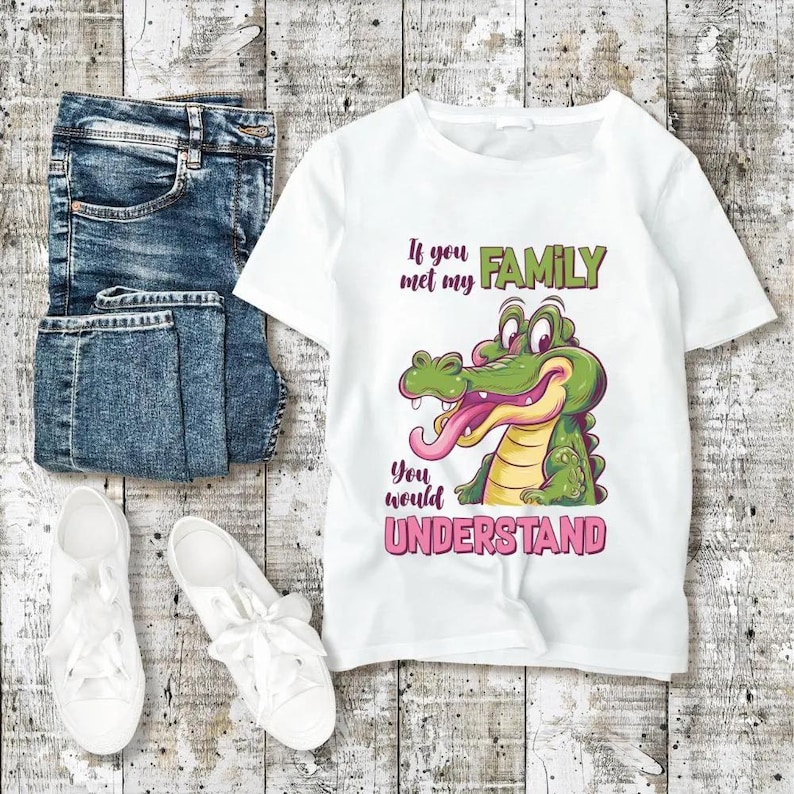 May include: White t-shirt with a cartoon alligator graphic and the text "If you met my FAMILY you would UNDERSTAND." The shirt is paired with blue jeans and white sneakers on a wooden surface.