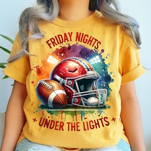 Puede incluir: Una camiseta de color amarillo mostaza con un gr&aacute;fico de un casco de f&uacute;tbol americano rojo y un bal&oacute;n de f&uacute;tbol marr&oacute;n. El dise&ntilde;o incluye un fondo de estilo acuarela colorido con luces de estadio y el texto FRIDAY NIGHTS UNDER THE LIGHTS en rojo.
