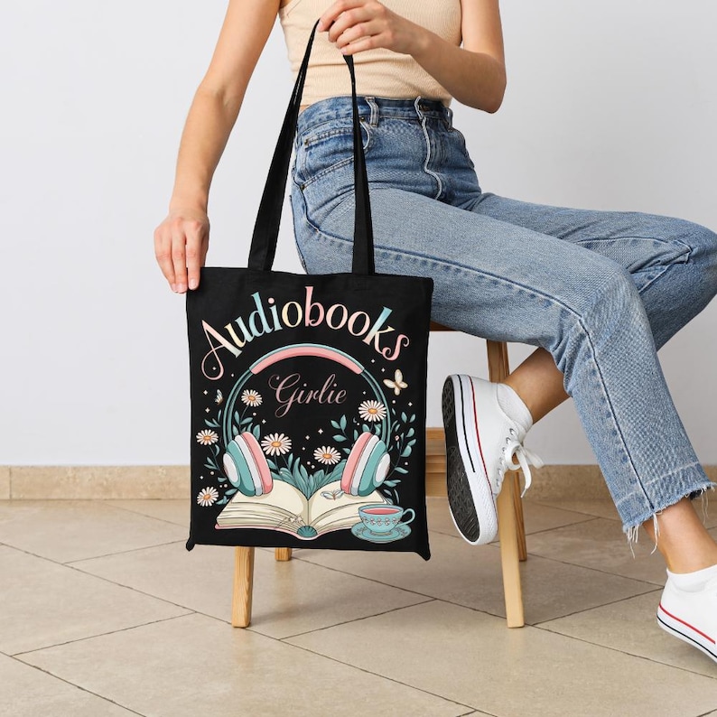 Puede incluir: Bolso tote negro con las palabras "Audiobooks Girlie" y un dise&ntilde;o de auriculares, flores, un libro abierto y una taza de t&eacute;. El bolso tiene asas negras. El dise&ntilde;o es en colores pastel.