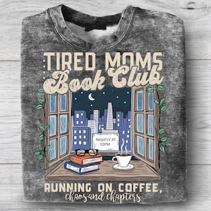 Puede incluir: Sudadera gris con un dise&ntilde;o gr&aacute;fico que muestra un horizonte urbano nocturno. El texto dice "TIRED MOMS Book Club" y "RUNNING ON COFFEE, chaos and chapters". Una taza de caf&eacute;, libros y gafas est&aacute;n en el alf&eacute;izar de la ventana.
