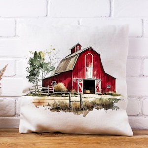 May include: A decorative pillow featuring a watercolor painting of a red barn with a white door and roof. The barn is set against a backdrop of a wooden fence, trees, and grassy field. The pillow is on a wooden surface.