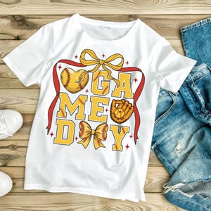 May include: White t-shirt with a "GAME DAY" design in yellow, featuring a baseball heart, a glove, and bows. The design is framed by a red ribbon. The shirt is paired with blue jeans and white sneakers.