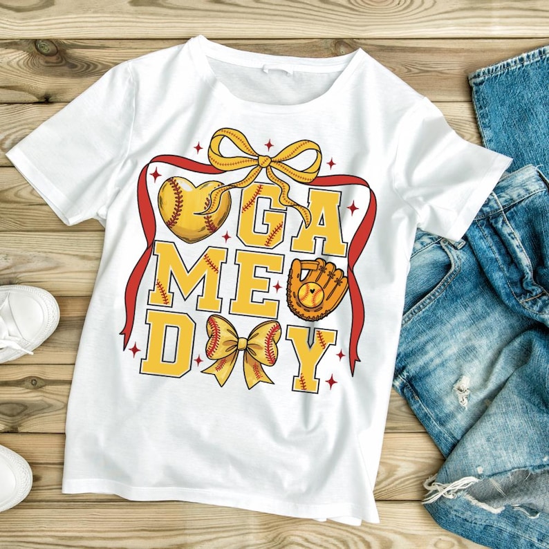 May include: White t-shirt with a "GAME DAY" design in yellow, featuring a baseball heart, a glove, and bows. The design is framed by a red ribbon. The shirt is paired with blue jeans and white sneakers.