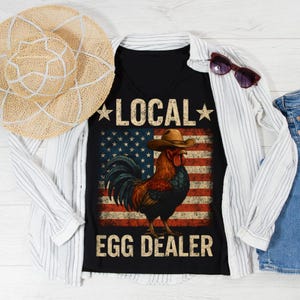 May include: Black V-neck t-shirt with a graphic of a rooster wearing a cowboy hat in front of an American flag. The text reads "LOCAL" above and "EGG DEALER" below the rooster. A straw hat and sunglasses are also in the image.