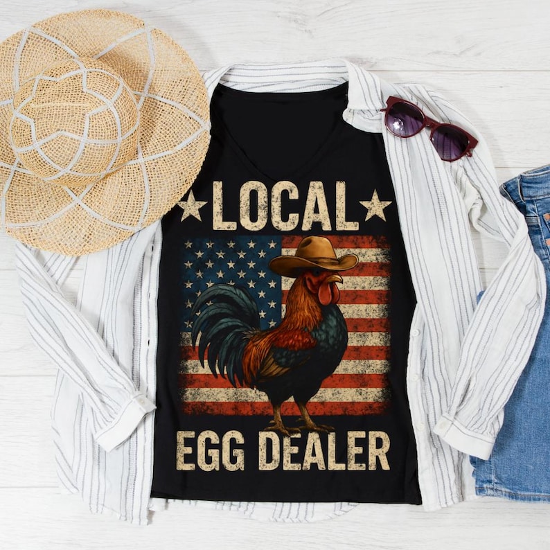 May include: Black V-neck t-shirt with a graphic of a rooster wearing a cowboy hat in front of an American flag. The text reads "LOCAL" above and "EGG DEALER" below the rooster. A straw hat and sunglasses are also in the image.