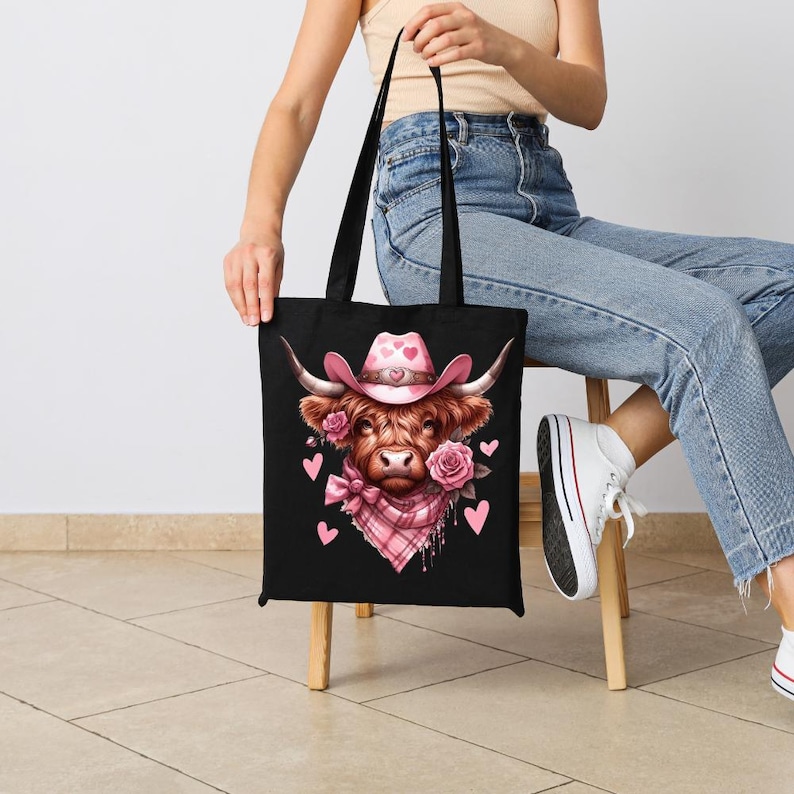 May include: Black tote bag featuring a watercolor illustration of a Highland cow wearing a pink cowboy hat with hearts. The cow has pink roses and a pink plaid scarf. The bag has black handles.