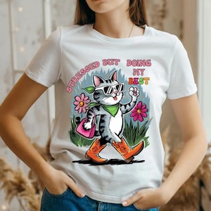 May include: White t-shirt featuring a cartoon cat wearing sunglasses, a green bandana, and orange cowboy boots. The cat is carrying a pink purse and walking past pink flowers and green grass. The text on the shirt reads "STRESSED BUT DOING MY BEST."