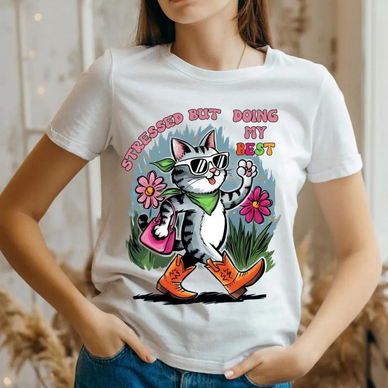 May include: White t-shirt featuring a cartoon cat wearing sunglasses, a green bandana, and orange cowboy boots. The cat is carrying a pink purse and walking past pink flowers and green grass. The text on the shirt reads "STRESSED BUT DOING MY BEST."