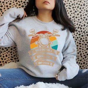 May include: A light gray sweatshirt with a motorcycle graphic and the text "Biker Dad Like a Normal Dad Only Cooler" in orange and white. The graphic features a motorcycle and a sunset design.