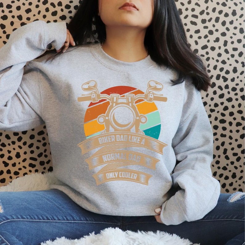 May include: A light gray sweatshirt with a motorcycle graphic and the text "Biker Dad Like a Normal Dad Only Cooler" in orange and white. The graphic features a motorcycle and a sunset design.