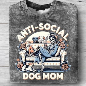 May include: Gray sweatshirt with the words "ANTI-SOCIAL DOG MOM" in a whimsical design. The graphic features a skeleton lounging on a couch with dogs, roses, and a coffee cup. The sweatshirt is folded.