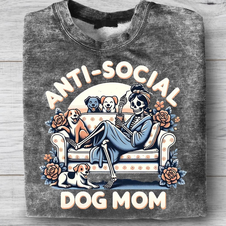 May include: Gray sweatshirt with the words "ANTI-SOCIAL DOG MOM" in a whimsical design. The graphic features a skeleton lounging on a couch with dogs, roses, and a coffee cup. The sweatshirt is folded.