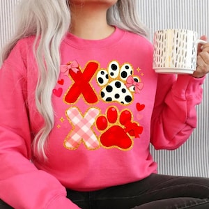 May include: A pink sweatshirt featuring a graphic design with the letters "X" and paw prints. The "X" letters are red and pink plaid, outlined in gold glitter. The paw prints are red and black and white, with pink bows and hearts.