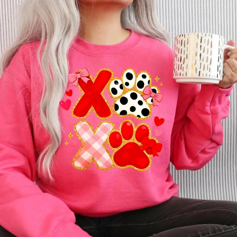 May include: A pink sweatshirt featuring a graphic design with the letters "X" and paw prints. The "X" letters are red and pink plaid, outlined in gold glitter. The paw prints are red and black and white, with pink bows and hearts.