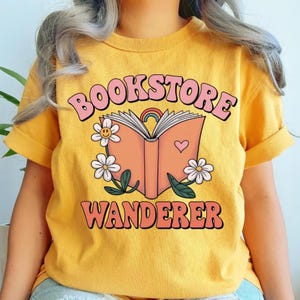 Puede incluir: Camiseta de color amarillo mostaza con las palabras "BOOKSTORE WANDERER" en una fuente retro. El dise&ntilde;o presenta un libro abierto con un arco&iacute;ris, un coraz&oacute;n y margaritas. La camiseta tiene las mangas enrolladas.