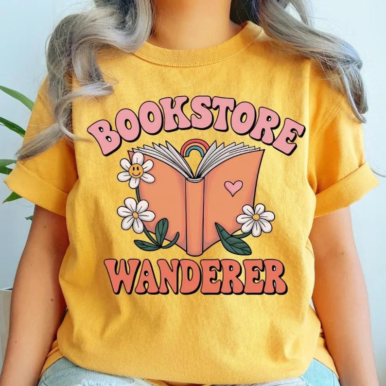 Puede incluir: Camiseta de color amarillo mostaza con las palabras "BOOKSTORE WANDERER" en una fuente retro. El dise&ntilde;o presenta un libro abierto con un arco&iacute;ris, un coraz&oacute;n y margaritas. La camiseta tiene las mangas enrolladas.