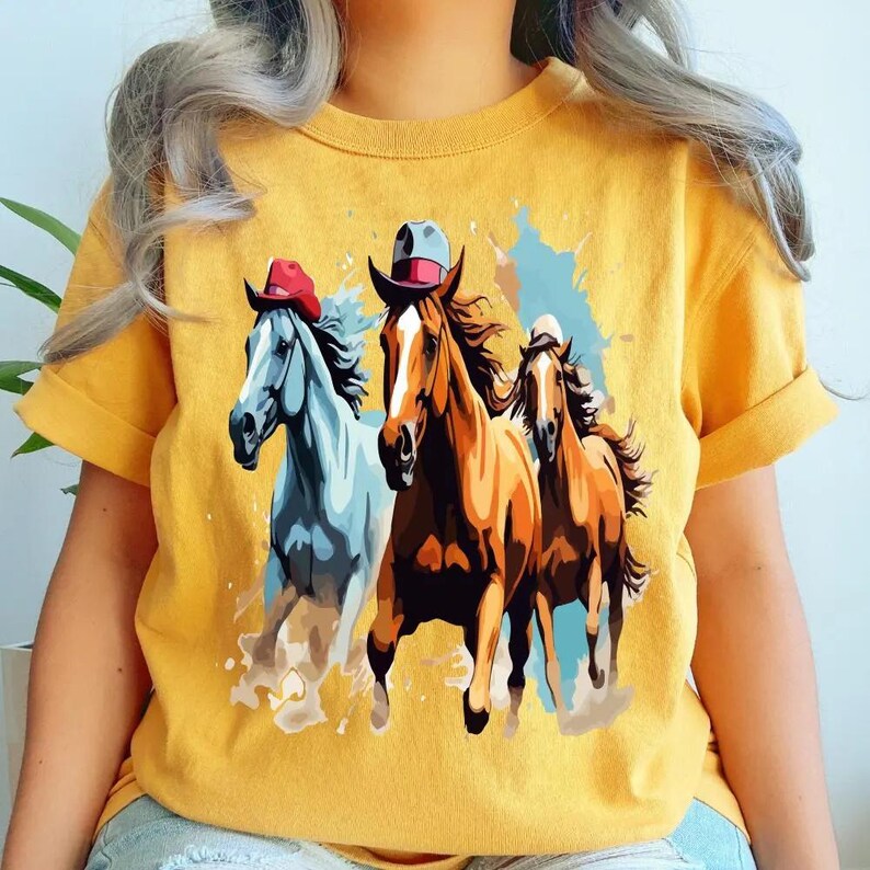 May include: Mustard yellow t-shirt featuring a graphic of three horses wearing cowboy hats. The horses are in shades of brown, blue, and white, with colorful paint splashes in the background. The shirt has short sleeves.