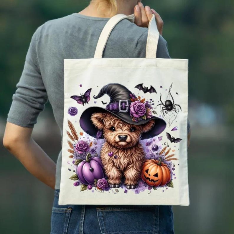 May include: A cream-colored tote bag featuring a whimsical Halloween design. The illustration includes a fluffy brown Highland cow wearing a witch hat, surrounded by purple roses, pumpkins, bats, a spider, and a butterfly. The bag has cream-colored handles.