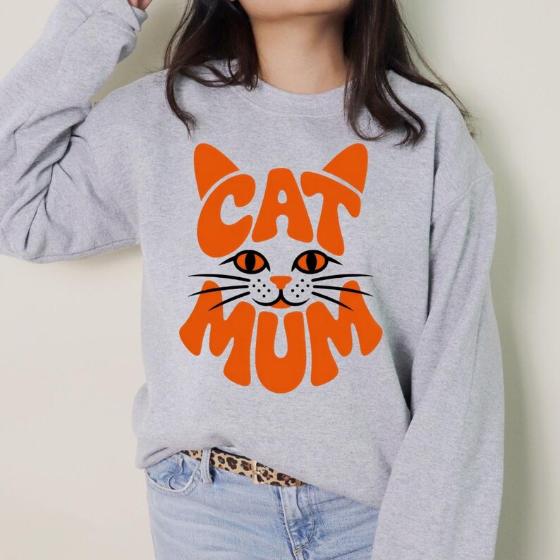 K&ouml;nnte beinhalten: Ein hellgraues Sweatshirt mit einem orangefarbenen Katzengesicht-Design. Das Design enth&auml;lt die W&ouml;rter "CAT" und "MUM" in Orange, die den Kopf und den K&ouml;rper der Katze bilden. Die Augen und Schnurrhaare der Katze sind schwarz.
