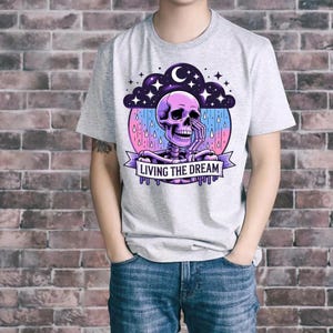 May include: A light gray t-shirt featuring a colorful graphic of a skeleton with a purple skull, a skeleton body, and the text "LIVING THE DREAM" on a banner. The design includes a night sky with stars and a crescent moon.