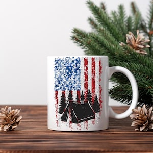 May include: White ceramic mug featuring a distressed American flag design with a black tent and trees silhouette. The mug has a curved handle and is suitable for hot or cold beverages. The design incorporates red, white, and blue colors, evoking a camping theme.