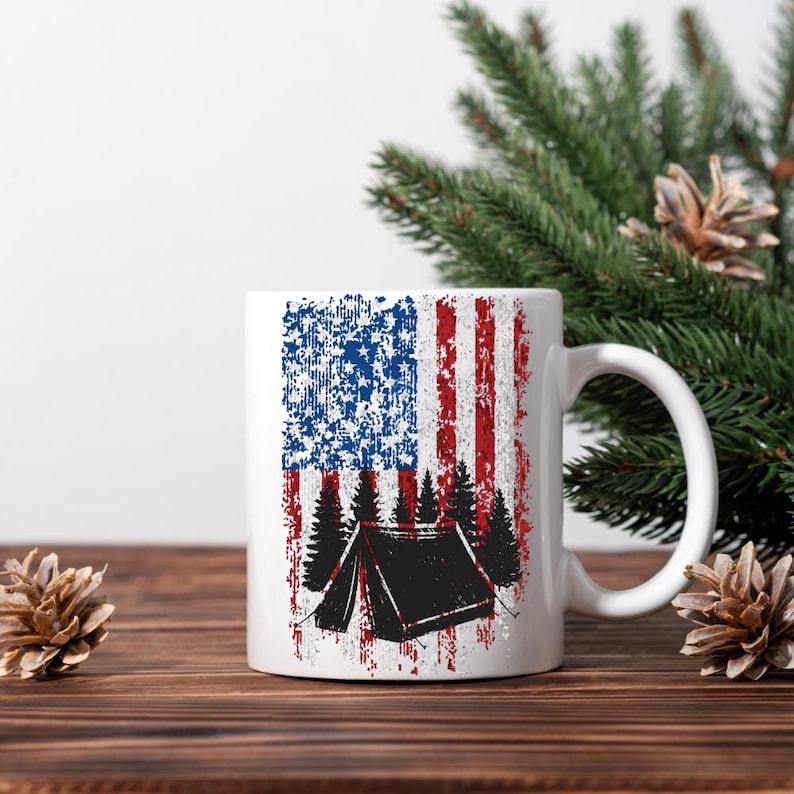 May include: White ceramic mug featuring a distressed American flag design with a black tent and trees silhouette. The mug has a curved handle and is suitable for hot or cold beverages. The design incorporates red, white, and blue colors, evoking a camping theme.