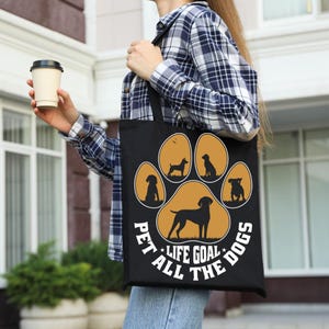 May include: Black tote bag featuring a paw print design with silhouettes of various dog breeds in a golden color. The text "PET ALL THE DOGS LIFE GOAL" is printed around the paw print. The bag is carried by a person wearing a plaid shirt.