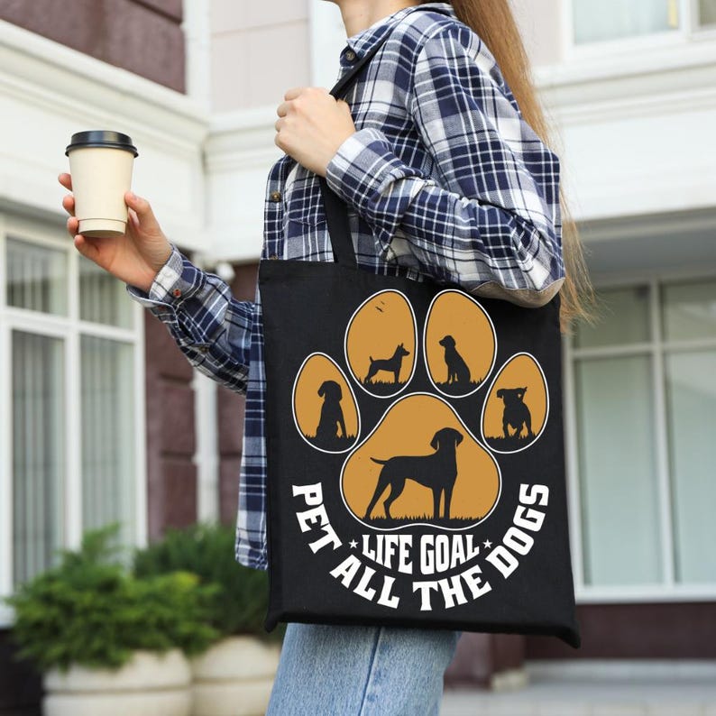 May include: Black tote bag featuring a paw print design with silhouettes of various dog breeds in a golden color. The text "PET ALL THE DOGS LIFE GOAL" is printed around the paw print. The bag is carried by a person wearing a plaid shirt.