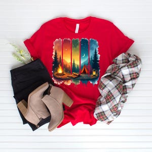 May include: A red t-shirt featuring a camping scene with a campfire, tent, and trees. The graphic has a colorful design with shades of orange, blue, and green. The shirt is paired with black pants, beige boots, and a plaid scarf.