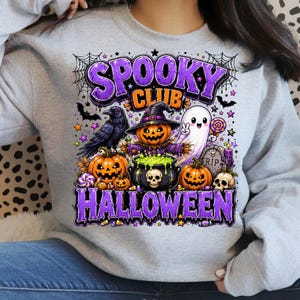 May include: A light gray sweatshirt featuring a graphic print with the text SPOOKY CLUB HALLOWEEN in purple. The design includes a black crow, a jack-o'-lantern wearing a witch hat, a ghost, pumpkins, a cauldron with green liquid, and a tombstone.