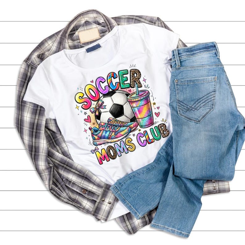 May include: White t-shirt with a colorful graphic that says "Soccer Moms Club". The graphic includes a soccer ball, sneakers, and a drink. The shirt is paired with blue jeans and a plaid shirt.