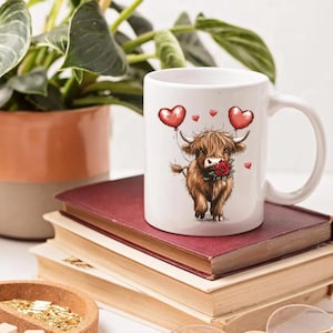 May include: White ceramic mug featuring a cartoon Highland cow holding a red rose and heart-shaped balloons. The cow is brown with long, shaggy fur. The mug is sitting on a stack of books, with a plant in the background.