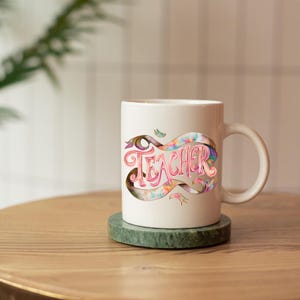 May include: White ceramic mug with the word "Teacher" in pink script, set against a colorful ribbon design. The mug sits on a green coaster, with a blurred background of a wooden table and greenery.