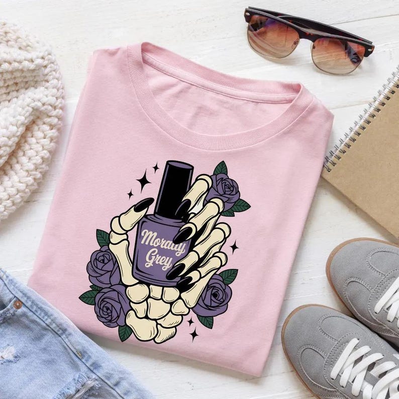 Puede incluir: Camiseta rosa claro con un dise&ntilde;o gr&aacute;fico. El dise&ntilde;o presenta una mano de esqueleto sosteniendo un frasco de esmalte de u&ntilde;as morado, con las palabras "Moray Grey". Tambi&eacute;n se incluyen rosas moradas y estrellas negras.