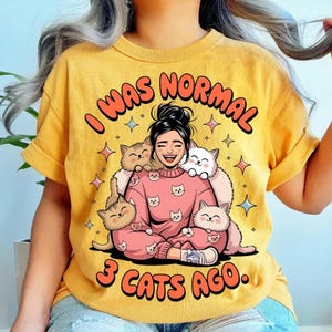 May include: Mustard yellow t-shirt with the text "I WAS NORMAL 3 CATS AGO." The graphic shows a smiling person surrounded by cartoon cats, wearing a pink sweater with cat faces. The shirt has rolled-up sleeves.