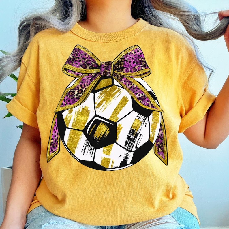 May include: Mustard yellow t-shirt featuring a soccer ball design with gold brushstrokes and black accents. A purple and gold leopard print bow is tied around the soccer ball. The shirt has rolled-up sleeves.