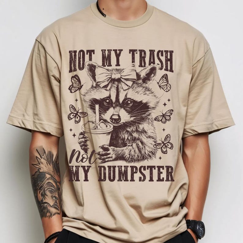 May include: Tan t-shirt featuring a raccoon with a bow, holding a drink, and surrounded by butterflies. The text reads "NOT MY TRASH, NOT MY DUMPSTER" in a brown, vintage font. The shirt is a casual, unisex style.