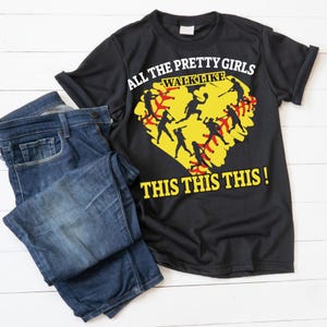 May include: Black t-shirt with the text "ALL THE PRETTY GIRLS WALK LIKE THIS THIS THIS!" and a yellow softball graphic with black silhouettes of softball players. The shirt is paired with blue denim jeans.