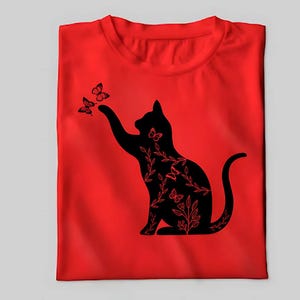 Pode incluir: Uma camiseta vermelha com uma silhueta preta de um gato com detalhes florais e borboletas. O gato est&aacute; alcan&ccedil;ando as borboletas.
