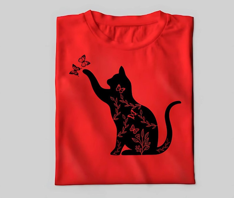 Pode incluir: Uma camiseta vermelha com uma silhueta preta de um gato com detalhes florais e borboletas. O gato est&aacute; alcan&ccedil;ando as borboletas.