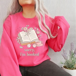 Puede incluir: Sudadera rosa brillante con un gr&aacute;fico de un libro abierto, una taza de t&eacute; y gafas. El texto "sorry, I'm booked" est&aacute; debajo del gr&aacute;fico. El dise&ntilde;o est&aacute; rodeado de flores y hojas.