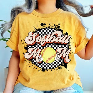 May include: Mustard yellow t-shirt with the words "Softball Mom" in a retro font. The design includes a softball and peace signs. The shirt has a black and white checkered background and black paint splatters.