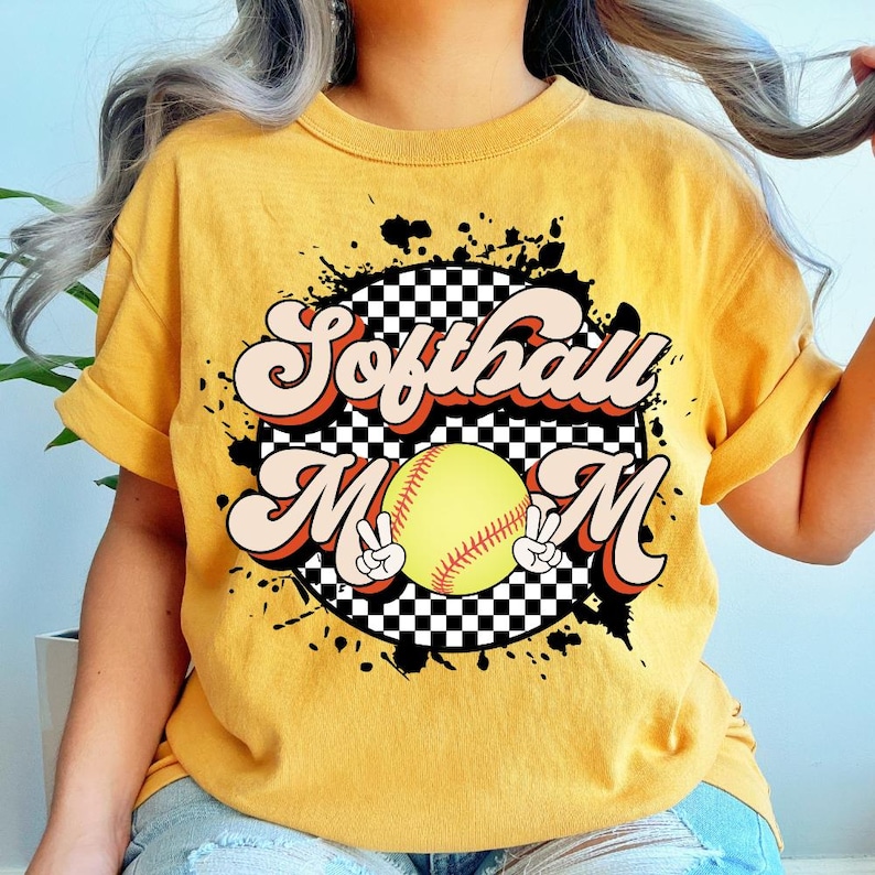 May include: Mustard yellow t-shirt with the words "Softball Mom" in a retro font. The design includes a softball and peace signs. The shirt has a black and white checkered background and black paint splatters.