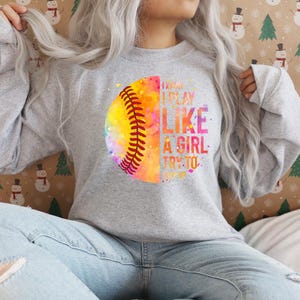 May include: A light gray sweatshirt featuring a colorful softball design. The softball is split, with a watercolor effect on one side and the other side displaying the text "I know I play like a girl try to keep up".