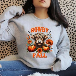 May include: A light gray sweatshirt featuring a graphic of a Highland cow with the words "HOWDY FALL" in orange. The design includes pumpkins, flowers, and foliage, perfect for autumn.