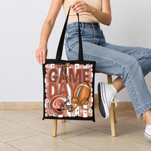 Puede incluir: Un bolso tote negro con un estampado gr&aacute;fico que incluye el texto GAME DAY en letras grandes de color marr&oacute;n. El dise&ntilde;o presenta un casco de f&uacute;tbol americano marr&oacute;n, un bal&oacute;n de f&uacute;tbol con un lazo y flores blancas sobre rayas verticales.